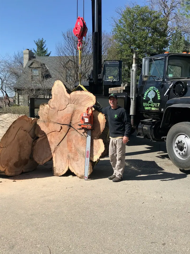 Smiling crew member stands by a massive tree trunk section with a large chainsaw and crane truck