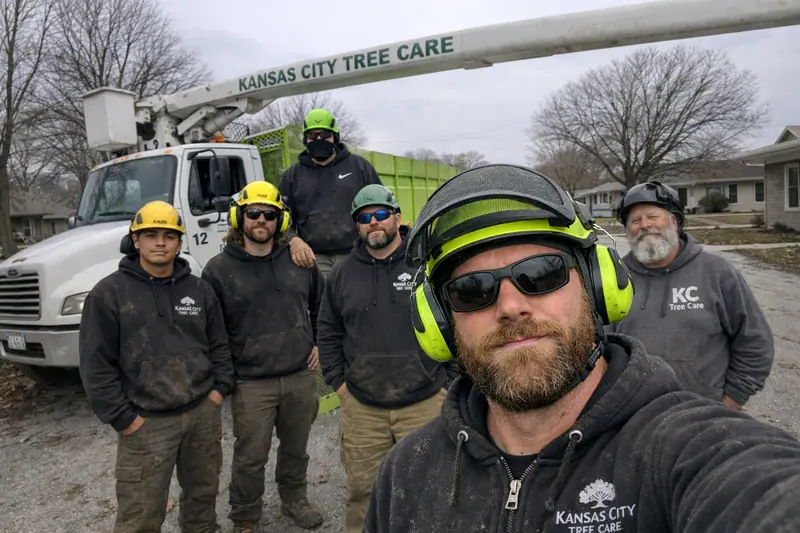 Six tree care crew members in hard hats pose with a Kansas City Tree Care bucket truck and chipper