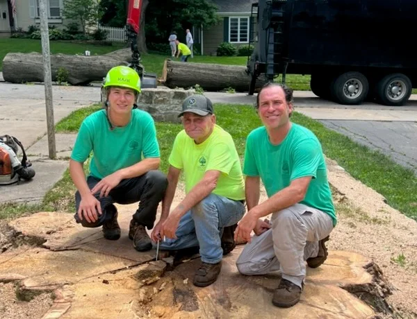 Zach Johnson kneeling on a large tree stump with his dad and his son, a Kansas City Tree Care job behind them