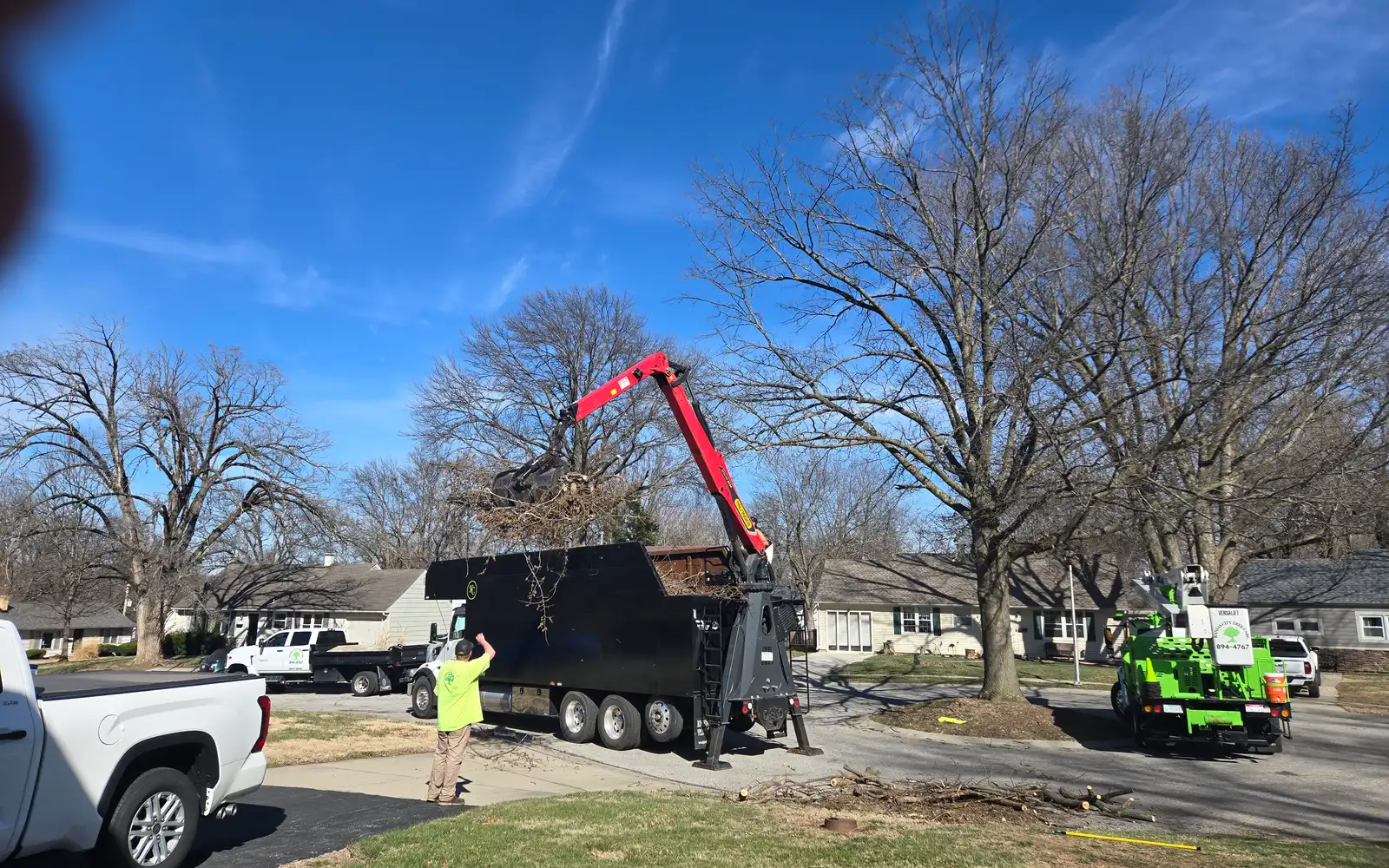 Kansas City Tree Care grapple truck with a red boom loading brush on a residential street while a crew member guides it