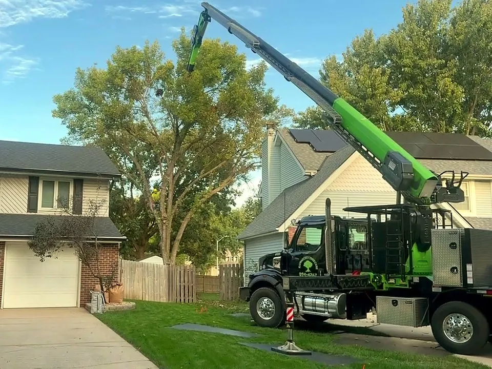 Kansas City Tree Care crane truck with its lime-green boom raised beside a two-story house