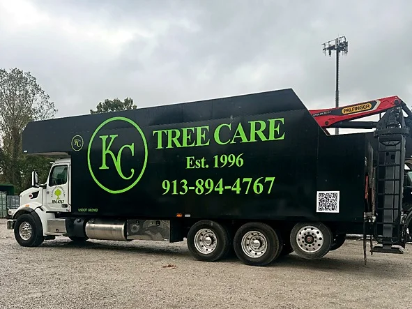 Black Kansas City Tree Care grapple truck with a red Palfinger loader crane behind the box, lettered KC Tree Care, Est. 1996, 913-894-4767